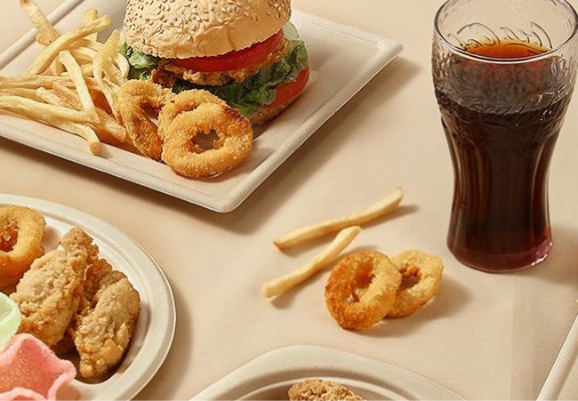 Fast food items served on bamboo fiber plates, including a burger, fries, onion rings, chicken, and a drink