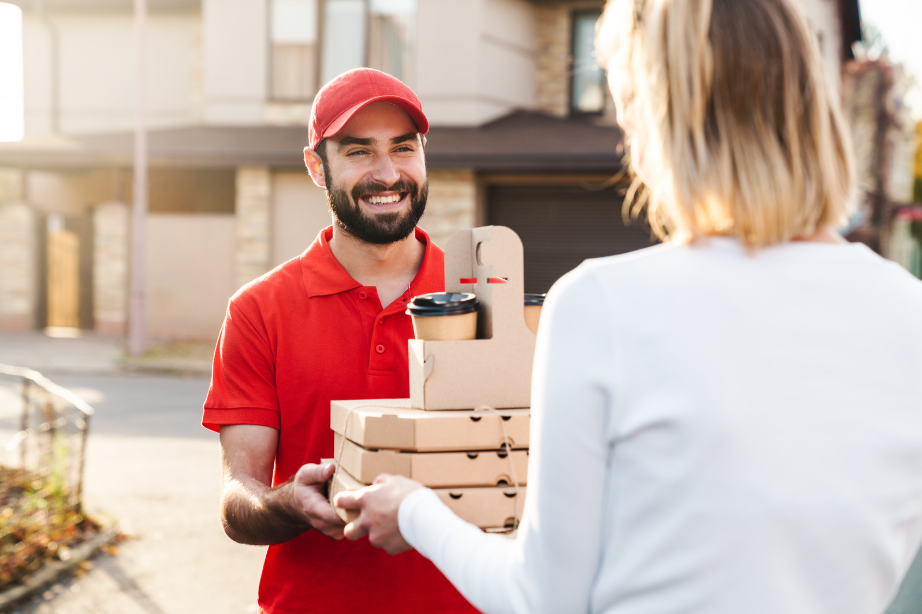 A delivery person in a red uniform handing over pizza boxes and coffee cups to a customer at their doorstep, representing a food delivery service.