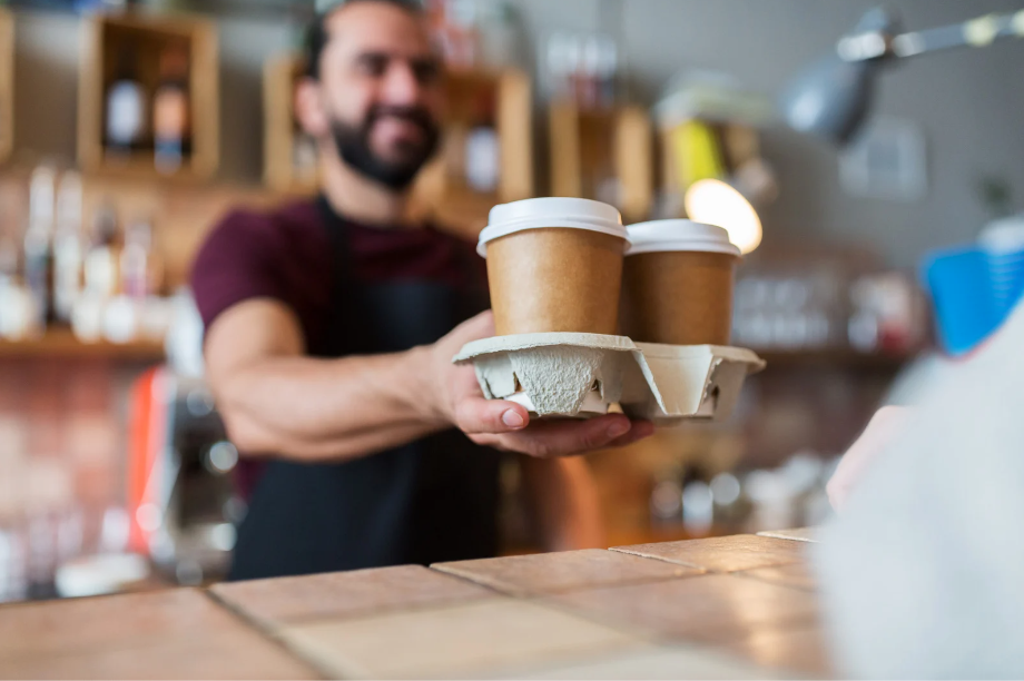 A barista handing over two eco-friendly coffee cups in a kraft paper design, placed in a cardboard carrier, representing sustainable coffee service.