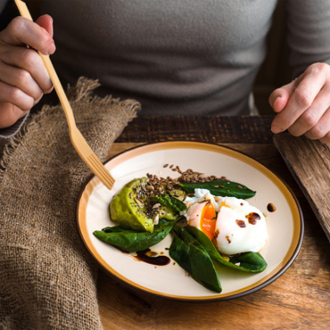 A person using a bamboo fork to eat a meal with poached eggs, avocado, and spinach on a rustic wooden table.