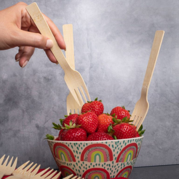 A hand picking up a biodegradable bamboo fork from a bowl of fresh strawberries, with multiple bamboo forks inserted.