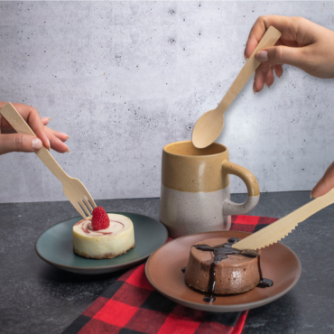 Hands using bamboo cutlery—fork, knife, and spoon—to enjoy cheesecake, chocolate dessert, and a hot beverage.